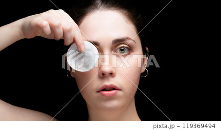 Beautiful young woman holding a cotton pad near her eye. Black background with copy space Beautiful young woman holding a cotton pad near her eye. Black background with copy space 119369494