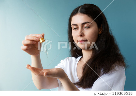 A young pretty Caucasian woman with dark hair drips a cosmetic product into the palm of her hand with a pipette. Blue background. The concept of hair care A young pretty Caucasian woman with dark hair drips a cosmetic product into the palm of her hand with a pipette. Blue background. The concept of hair care 119369519