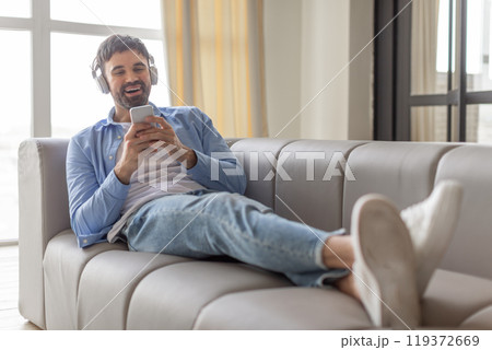 A man is lying on a grey couch with his legs extended and wearing headphones. He is smiling and looking at his phone. 119372669