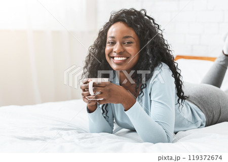Young african woman enjoying aromatic coffee in bed, smiling at camera, free space 119372674