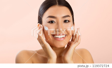 Asian young woman with dark hair applying white face cream to her cheeks, smiling at the camera. She has a fresh, clean complexion and appears to be enjoying the process of applying the cream Asian young woman with dark hair applying white face cream to her cheeks, smiling at the camera. She has a fresh, clean complexion and appears to be enjoying the process of applying the cream 119372724