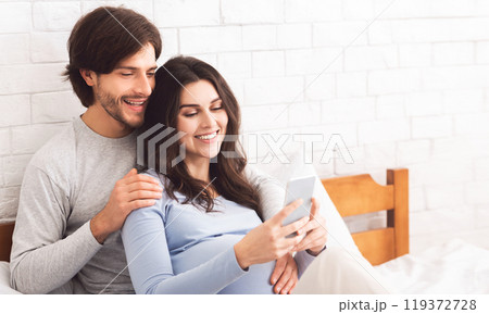 Happy pregnant couple posing for selfie, cuddling in bed at home, enjoying lazy weekend together, free space 119372728