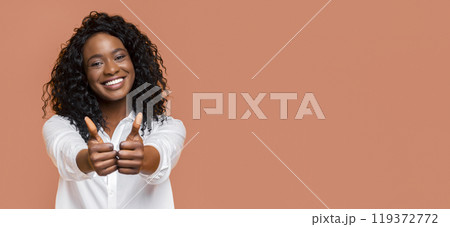 A young black woman with curly black hair smiles brightly at the camera, giving two thumbs up with both hands extended. She is wearing a white button-down shirt and standing against peach background. 119372772