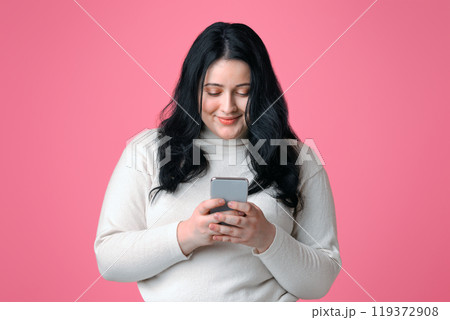 Smiling plump woman holding smartphone in hands, texting, reading message, or using new application, isolated on pink background with free space Smiling plump woman holding smartphone in hands, texting, reading message, or using new application, isolated on pink background with free space 119372908