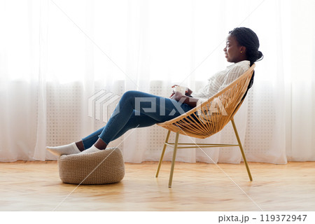 Relax at home. African american girl resting in wicker armchair and drinking coffee against window, free space 119372947