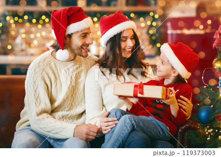 Beautiful smiling parents in santa hats giving xmas gift to their daughter, celebrating Christmas at home, copy space Beautiful smiling parents in santa hats giving xmas gift to their daughter, celebrating Christmas at home, copy space 119373003