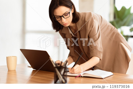 Self employed female entrepreneur is busy writing in her notebook, standing at desk with large window in the background 119373016