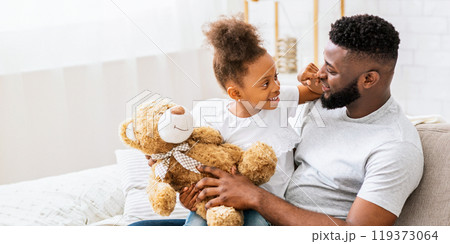 African American father and daughter are sitting on a couch together at home. They are both smiling and looking at each other. The daughter is holding a teddy bear. African American father and daughter are sitting on a couch together at home. They are both smiling and looking at each other. The daughter is holding a teddy bear. 119373064