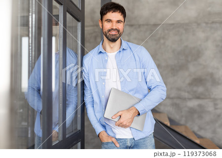 A man in a blue shirt and jeans stands in an office space, smiling and holding a laptop in his hands. He is looking directly at the camera, and his left hand is in his pocket. A man in a blue shirt and jeans stands in an office space, smiling and holding a laptop in his hands. He is looking directly at the camera, and his left hand is in his pocket. 119373068