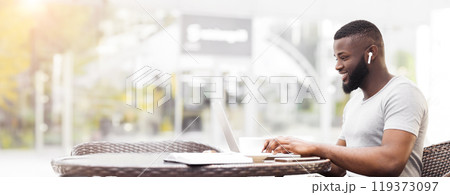 A man sits at an outdoor cafe table, working on his laptop. He is smiling and wearing headphones, suggesting he is enjoying his work. A man sits at an outdoor cafe table, working on his laptop. He is smiling and wearing headphones, suggesting he is enjoying his work. 119373097
