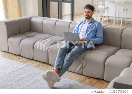 A man sits on a grey modular sofa in a modern living room, using a laptop computer. He is smiling and looks relaxed, possibly enjoying a break from work, high angle view 119373103
