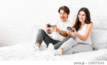 A couple sits on a white bed in a white room, playing video games with controllers in their hands. They both look excited and engaged in the game. A couple sits on a white bed in a white room, playing video games with controllers in their hands. They both look excited and engaged in the game. 119373143