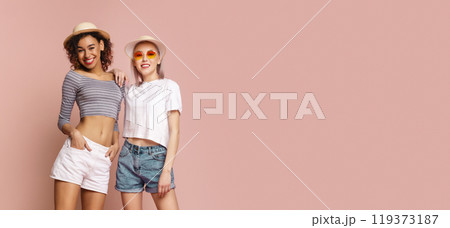 This image shows two young women standing in front of a pink background. Both women are wearing summer attire, including shorts, hats, and sunglasses, copy space This image shows two young women standing in front of a pink background. Both women are wearing summer attire, including shorts, hats, and sunglasses, copy space 119373187