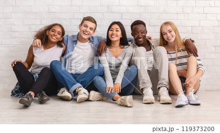 Multiracial happy students posing over white brick wall, sitting on floor and embracing, friendship concept 119373200