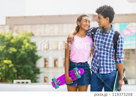 Teen black couple walking down the street in an urban area. Guy has his arm around the girl, and they are both smiling and looking at each other. They are carrying a skateboard, copy space Teen black couple walking down the street in an urban area. Guy has his arm around the girl, and they are both smiling and looking at each other. They are carrying a skateboard, copy space 119373208