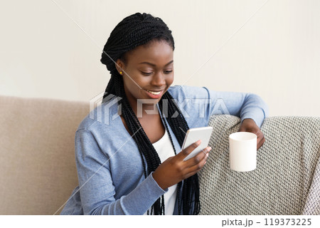 Leisure concept. Young african american woman texting on smartphone and drinking coffee, relaxing on comfortable sofa at home, free space 119373225