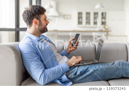 A man sits on a couch in a modern home, comfortably using a smartphone and a laptop. He is smiling, likely engaging with a digital task or enjoying some entertainment. A man sits on a couch in a modern home, comfortably using a smartphone and a laptop. He is smiling, likely engaging with a digital task or enjoying some entertainment. 119373243