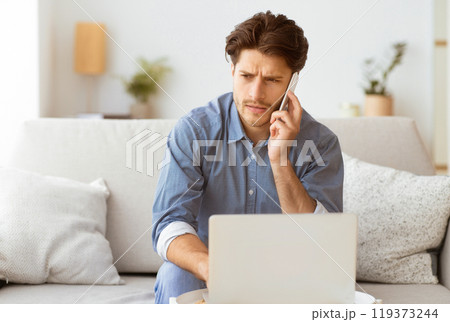 A young man sits on a couch, working on his laptop while talking on his phone. He appears to be focused and engaged in a conversation. A young man sits on a couch, working on his laptop while talking on his phone. He appears to be focused and engaged in a conversation. 119373244