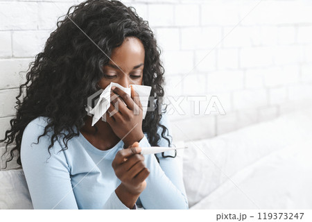 Sick african woman having fever, measuring body temperature with thermometer, blowing nose in bed, copy space 119373247