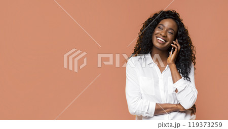 African American young woman with long curly hair is smiling while talking on a phone in front of a solid orange background. She is wearing a white shirt and has her arms crossed. 119373259