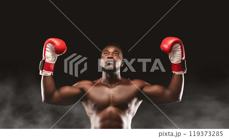 A muscular boxer celebrates a victory with arms raised, wearing red boxing gloves against a dark smoky background. 119373285