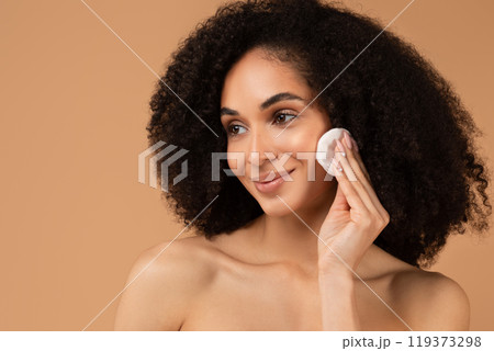 A young African American woman with curly hair smiles while applying makeup to her cheek with a cotton pad in her right hand. The background is a warm, light brown color. A young African American woman with curly hair smiles while applying makeup to her cheek with a cotton pad in her right hand. The background is a warm, light brown color. 119373298