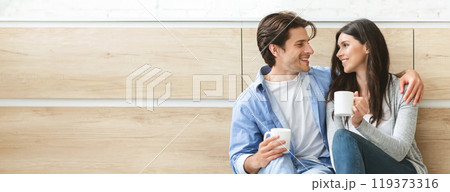 A couple is sitting on the floor in a kitchen, drinking coffee and talking. They both have relaxed expressions and are looking at each other lovingly, copy space 119373316