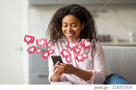 A young black woman with curly hair smiles as she uses her smartphone in her living room. Pink heart-shaped icons float around her, indicating social media likes and engagement. 119373335