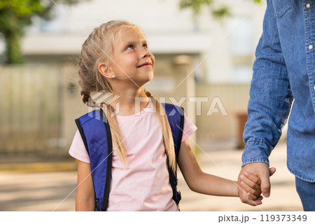 A young girl with blonde pigtails and a blue backpack walks to school holding the hand of her parent. She is looking up and smiling as they walk down the sidewalk. 119373349