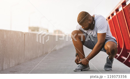 Black man tying running shoes on bridge, free space Black man tying running shoes on bridge, free space 119373356