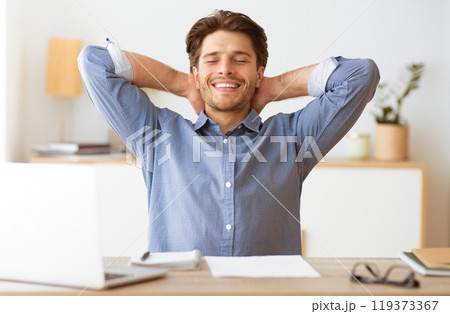 A relaxed man sits at his desk, leaning back with his arms behind his head, smiling and taking a break from work. He appears content and at ease. A relaxed man sits at his desk, leaning back with his arms behind his head, smiling and taking a break from work. He appears content and at ease. 119373367