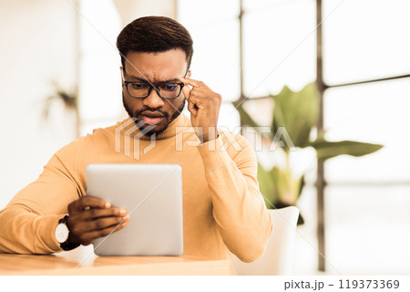 Bad News. Focused african american man looking at digital tablet at workplace, touching glasses, copyspace Bad News. Focused african american man looking at digital tablet at workplace, touching glasses, copyspace 119373369