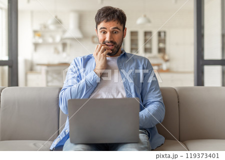 A man sits on a couch in his living room, biting his nails as he stares intently at a laptop in his lap. He looks concerned and apprehensive. 119373401