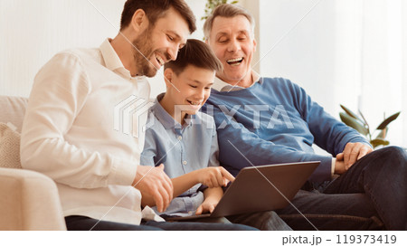Male Family Weekend. Joyful Boy Using Laptop Computer Sitting Between Father And Grandpa On Sofa At Home. Panorama 119373419