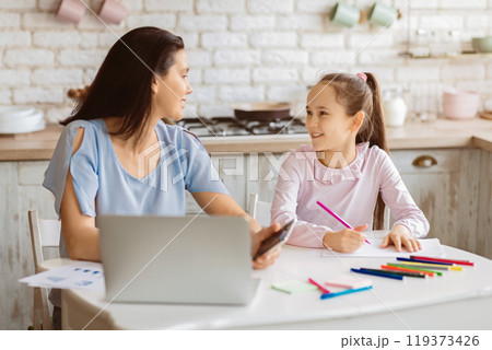 A woman works on her laptop while her daughter draws at the kitchen table. They are both smiling and appear happy to be spending time together. The kitchen is modern and bright 119373426