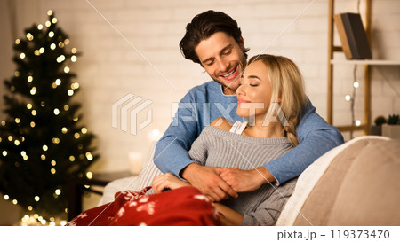 Cozy Christmas eve. Loving couple embracing near Christmas tree, resting on sofa 119373470