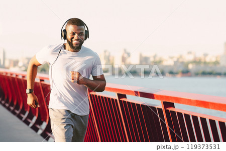 A young man smiles while jogging on a bridge with a city skyline in the background. He is wearing headphones and a white t-shirt. A young man smiles while jogging on a bridge with a city skyline in the background. He is wearing headphones and a white t-shirt. 119373531