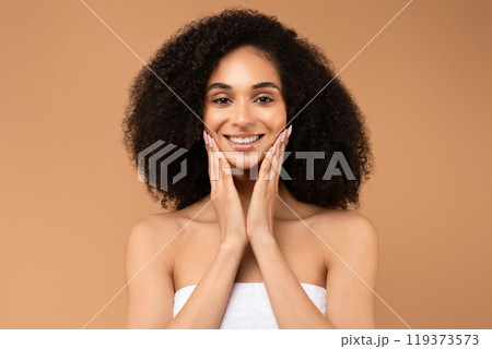 African American young woman with dark, curly hair smiles brightly as she holds her hands to her cheeks. She is wearing a white strapless top and standing in front of a plain, tan backdrop. 119373573