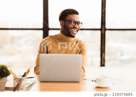 Success At Work. Happy afro businessman sitting at desk by large window in modern office, looking aside at copyspace 119373605
