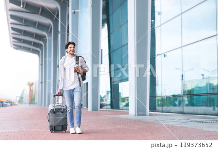 Going To Registration. Happy Young Man Walking With Suitcase And Backpack Near Airport Terminal, Holding Passport And Tickets In Hand, Free Space 119373652
