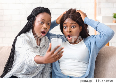 Unexpected Message. Two Shocked Black Girlfriends Looking At Smartphone Screen With Open Mouth, Sitting On Couch At Home Unexpected Message. Two Shocked Black Girlfriends Looking At Smartphone Screen With Open Mouth, Sitting On Couch At Home 119373656
