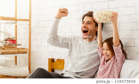 Cute emotional little girl and her father watching football match on TV, celebrating success of their team, sitting on bed, holding bowl with popcorn, family traditions concept Cute emotional little girl and her father watching football match on TV, celebrating success of their team, sitting on bed, holding bowl with popcorn, family traditions concept 119373658