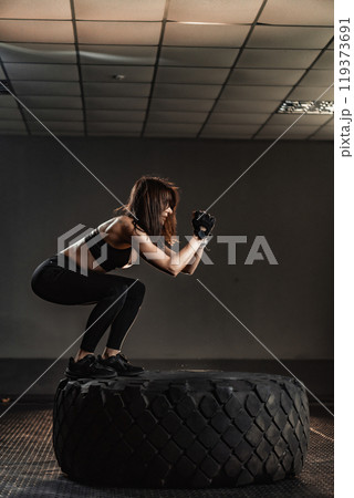 A dedicated female athlete focuses on tire squats to boost her strength and conditioning, improving her fitness for success in competition and preparing for intense training ahead A dedicated female athlete focuses on tire squats to boost her strength and conditioning, improving her fitness for success in competition and preparing for intense training ahead 119373691