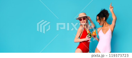 Two women in swimsuits are celebrating with drinks in their hands. They are standing in front of a bright blue background, panorama with copy space Two women in swimsuits are celebrating with drinks in their hands. They are standing in front of a bright blue background, panorama with copy space 119373723