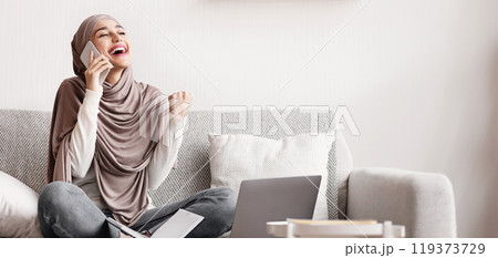 Good news on phone. Cheerful arabic woman in hijab talking on cellphone and celebrating success, she got job, sitiing on couch at home 119373729