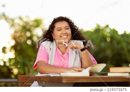 A young student sits at a wooden table surrounded by greenery, smiling while holding a pen and studying from an open book under the warm sunlight. A young student sits at a wooden table surrounded by greenery, smiling while holding a pen and studying from an open book under the warm sunlight. 119373875