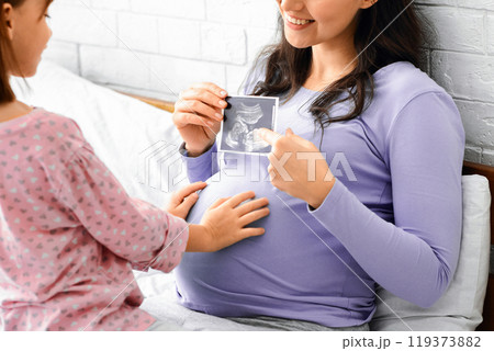Happy pregnant woman showing ultrasound scan of baby to her little daughter, sitting together on bed Happy pregnant woman showing ultrasound scan of baby to her little daughter, sitting together on bed 119373882
