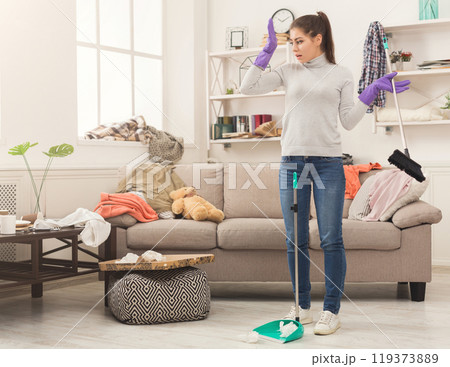 Shocked woman with special equipment cleaning house. Young girl surrounded by many stack of clothes. Disorder and mess at home, copy space 119373889
