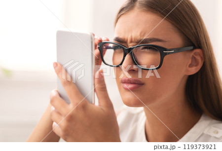 Poor Eyesight Concept. Girl Looking At Cellphone Screen Through Eyeglasses Having Vision Problem Indoor. Selective Focus Poor Eyesight Concept. Girl Looking At Cellphone Screen Through Eyeglasses Having Vision Problem Indoor. Selective Focus 119373921