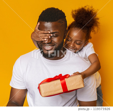 Father's day. Cheerful little afro daughter hugging her dad, closing his eyes and giving gift box, orange background 119373926